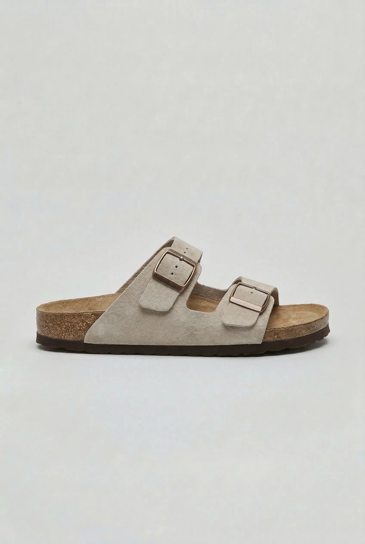Side profile of a light grey suede two-strap sandal with dark brown sole and metal buckles