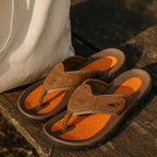 Pair of brown leather sandals with orange footbeds resting on a wooden bench beside a canvas bag