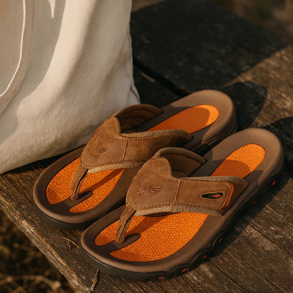 Pair of brown leather sandals with orange footbeds resting on a wooden bench beside a canvas bag