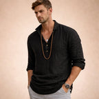 Man in a black linen popover shirt with three-button placket and classic collar, sleeves casually rolled up, worn with layered necklaces.
