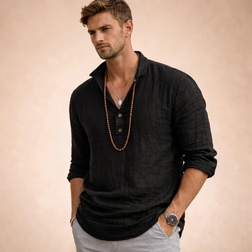 Man in a black linen popover shirt with three-button placket and classic collar, sleeves casually rolled up, worn with layered necklaces.