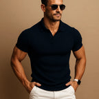 Man in a navy ribbed knit polo shirt with open collar, aviator sunglasses and white trousers, hands in pockets