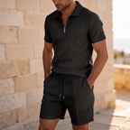 Man in a cream knit set with sunglasses, standing barefoot on a sunlit stone terrace