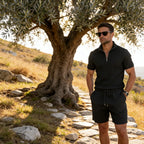 Man in a cream polo and shorts set gazing into the distance under olive branches on a hillside