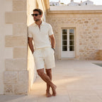 Side view of a man in a cream knit polo and shorts set leaning against a stone archway by a pool