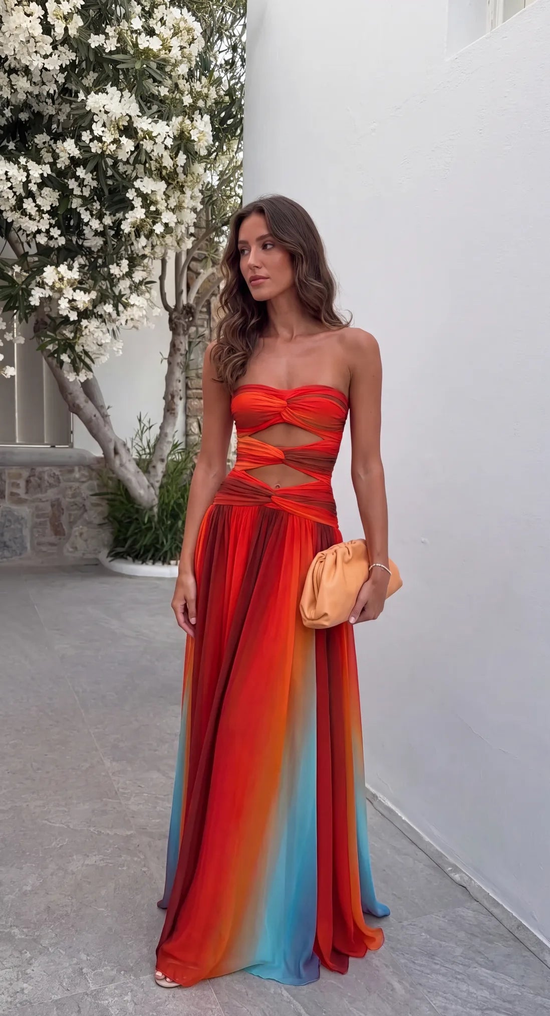 Strapless multicolour ombre maxi dress with front knot bodice and cutout waist, styled with a tan clutch bag beside flowering trees.