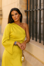 Model wearing a bright yellow one-shoulder maxi dress with long sleeve, styled with a gold chain handbag and hoop earrings