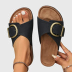 Black leather sandals with cork-look footbed and oversized antique gold buckle, shown from above on bare feet