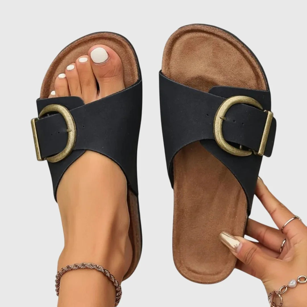 Black leather sandals with cork-look footbed and oversized antique gold buckle, shown from above on bare feet