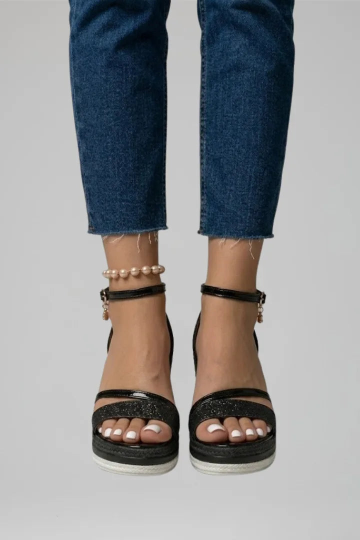 Close-up front view of feet wearing black glitter sandals with thin patent ankle straps and pearl anklet