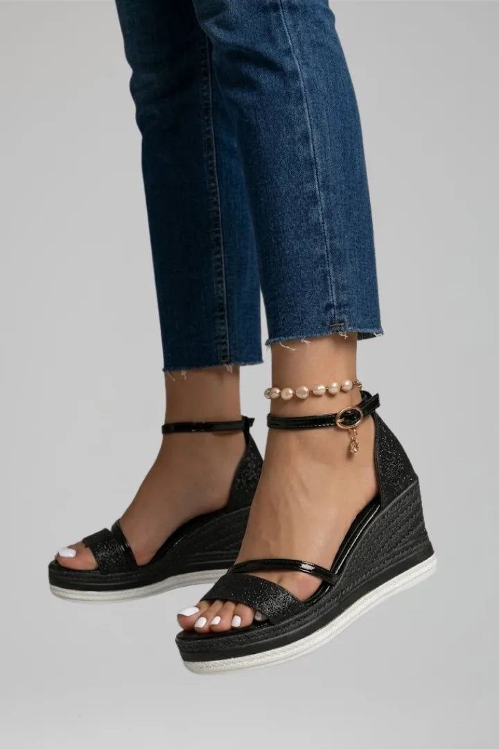 Close-up side view of black glitter wedge sandal in motion, highlighting ankle strap and charm detail