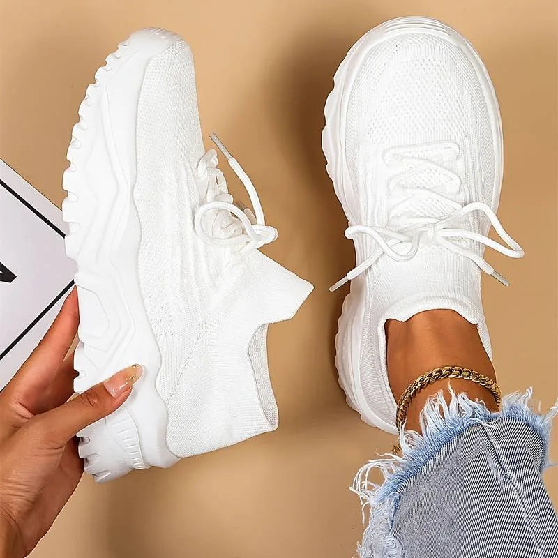 Overhead view of a hand holding a white knit sneaker beside its matching pair worn on foot, showing the chunky sole and sock-style ankle
