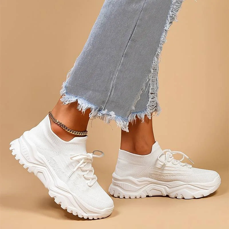 Side view of a white knit sneaker worn on foot with ripped denim jeans, showing the sock-like fit and lace-up front