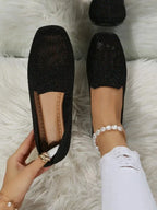 Black glittery knit loafers with perforated toe design, shown on bed beside a pearl anklet