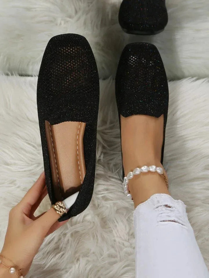 Black glittery knit loafers with perforated toe design, shown on bed beside a pearl anklet