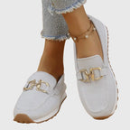 Close-up of beige knit loafers on crossed feet, highlighting the gold buckle and tan outsole
