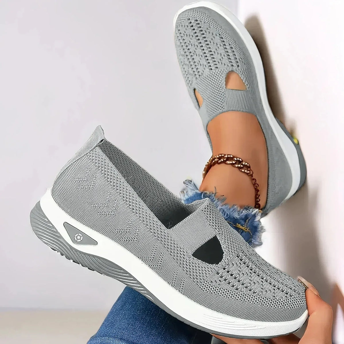 Close-up of grey knit slip-on shoe held in hand, showing textured woven upper, cut-out design and ridged white outsole
