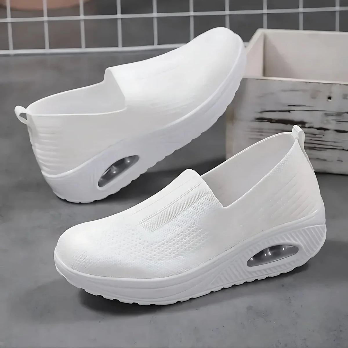 Pair of white knit slip-on sneakers displayed on concrete, showing air-cushion heel detail and textured sole
