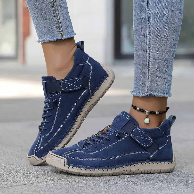 Navy blue suede sneaker with tan sole and hand-stitched panels, worn with cuffed denim jeans