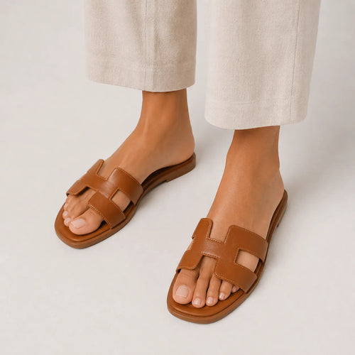 Close-up of feet wearing tan leather slide sandals with cut-out H strap, paired with cream wide-leg trousers