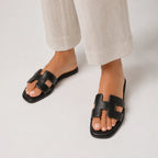 Feet in black leather slide sandals with H-strap cut-out detail, styled with cream cropped trousers