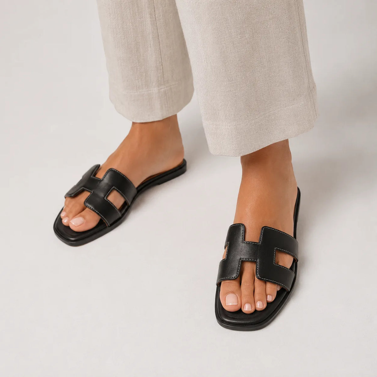 Feet in black leather slide sandals with H-strap cut-out detail, styled with cream cropped trousers