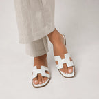Feet wearing white leather slide sandals with H-strap design, shown against light cream trousers