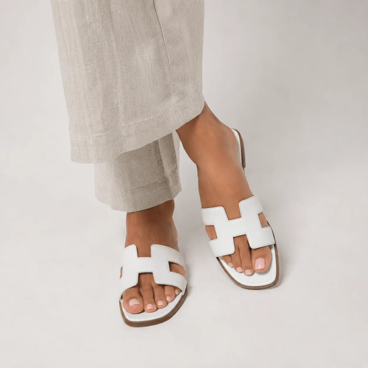 Feet wearing white leather slide sandals with H-strap design, shown against light cream trousers