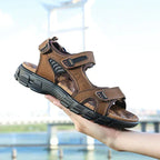 Hand holding a dark brown leather sandal with adjustable hook-and-loop straps and a chunky black outsole, bridge visible in the background.