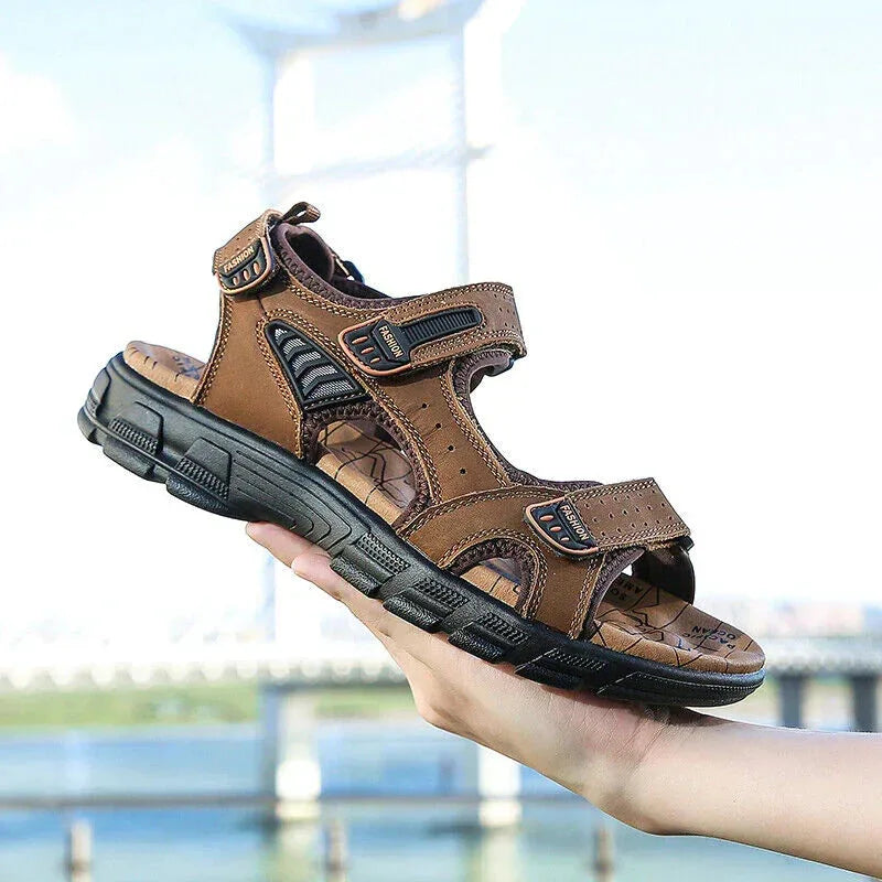 Hand holding a dark brown leather sandal with adjustable hook-and-loop straps and a chunky black outsole, bridge visible in the background.