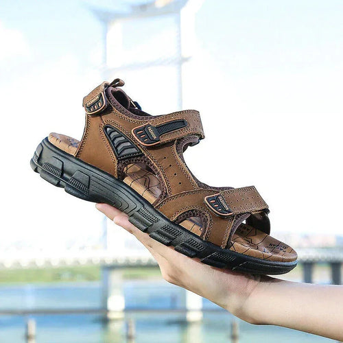Hand holding a dark brown leather sandal with adjustable hook-and-loop straps and a chunky black outsole, bridge visible in the background.