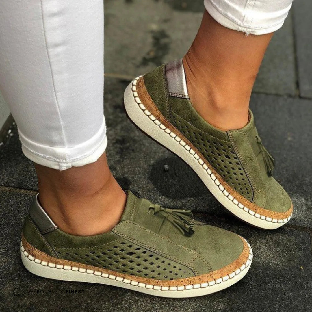 Side view of olive green perforated suede slip-on sneaker with cork-trim sole and tassel detail, worn with white jeans