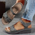 Grey wedge sandal with hook-and-loop ankle strap and toe strap, shown held up to display the cushioned tan footbed