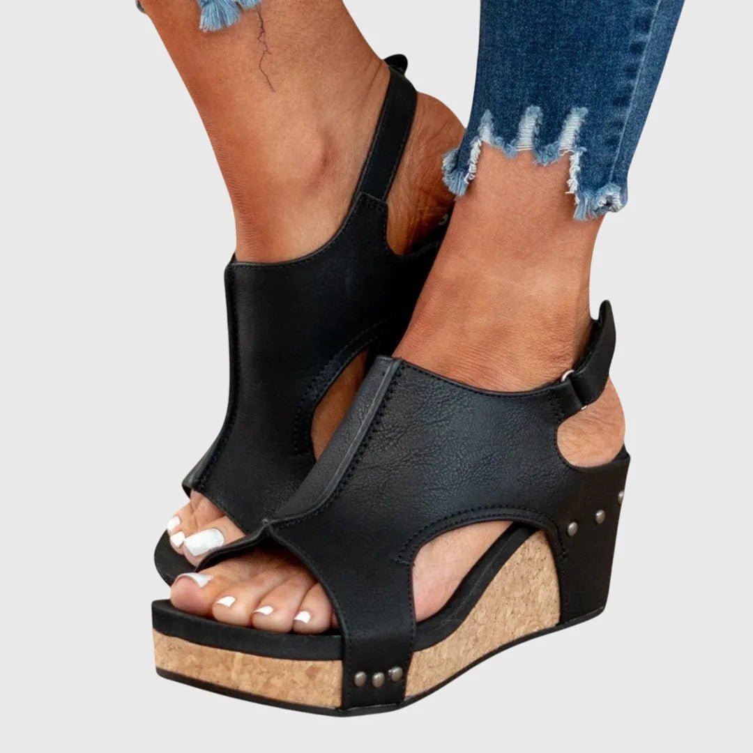 Front view of black wedge sandals with peep toe, shield vamp and cork platform sole worn with cuffed jeans
