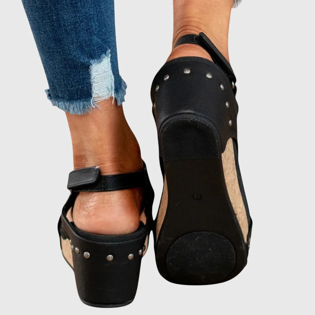 Close-up of black wedge sandals showing buckle ankle strap and stud trim along the cork wedge
