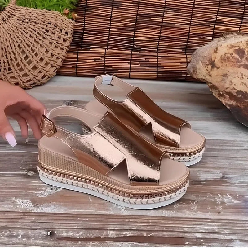 Pair of rose gold metallic wedge sandals resting on a timber surface showing studded platform trim