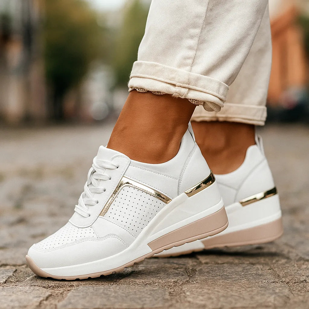 Close-up of white wedge sneakers with gold perforated panel and lace-up front, worn with cuffed cream trousers