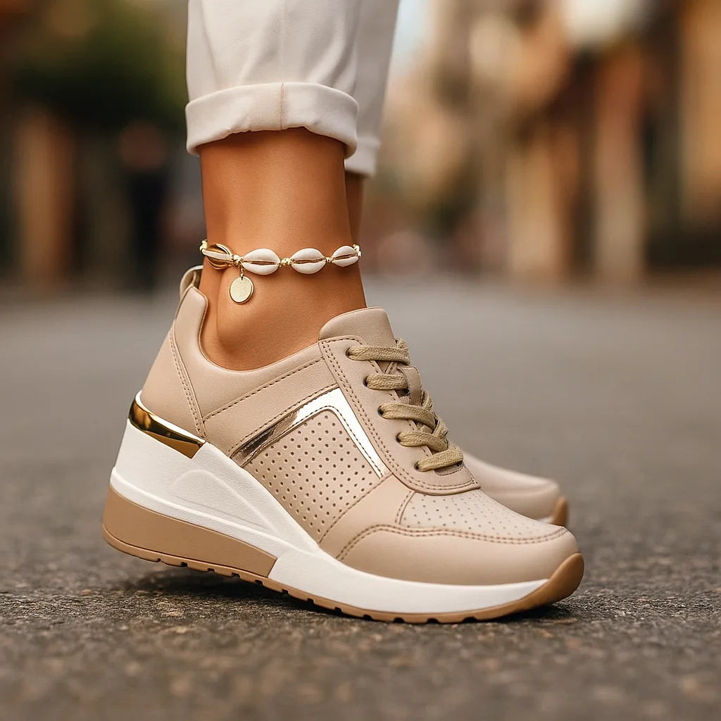 Beige wedge sneakers with gold heel trim and perforated side panel, shown close up with a shell anklet