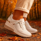 White wedge sneakers with gold accent strip, worn outdoors on a path scattered with autumn leaves