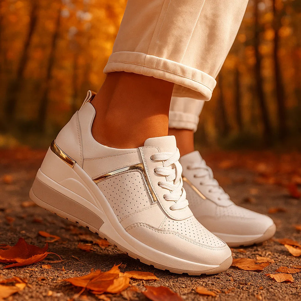 White wedge sneakers with gold accent strip, worn outdoors on a path scattered with autumn leaves