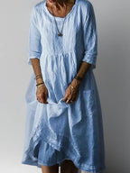 Light blue pleated linen midi dress with round neckline and three-quarter sleeves, worn with layered necklace