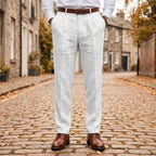 Front view of white pleated linen trousers worn with a brown leather belt and brogue shoes on a cobblestone street
