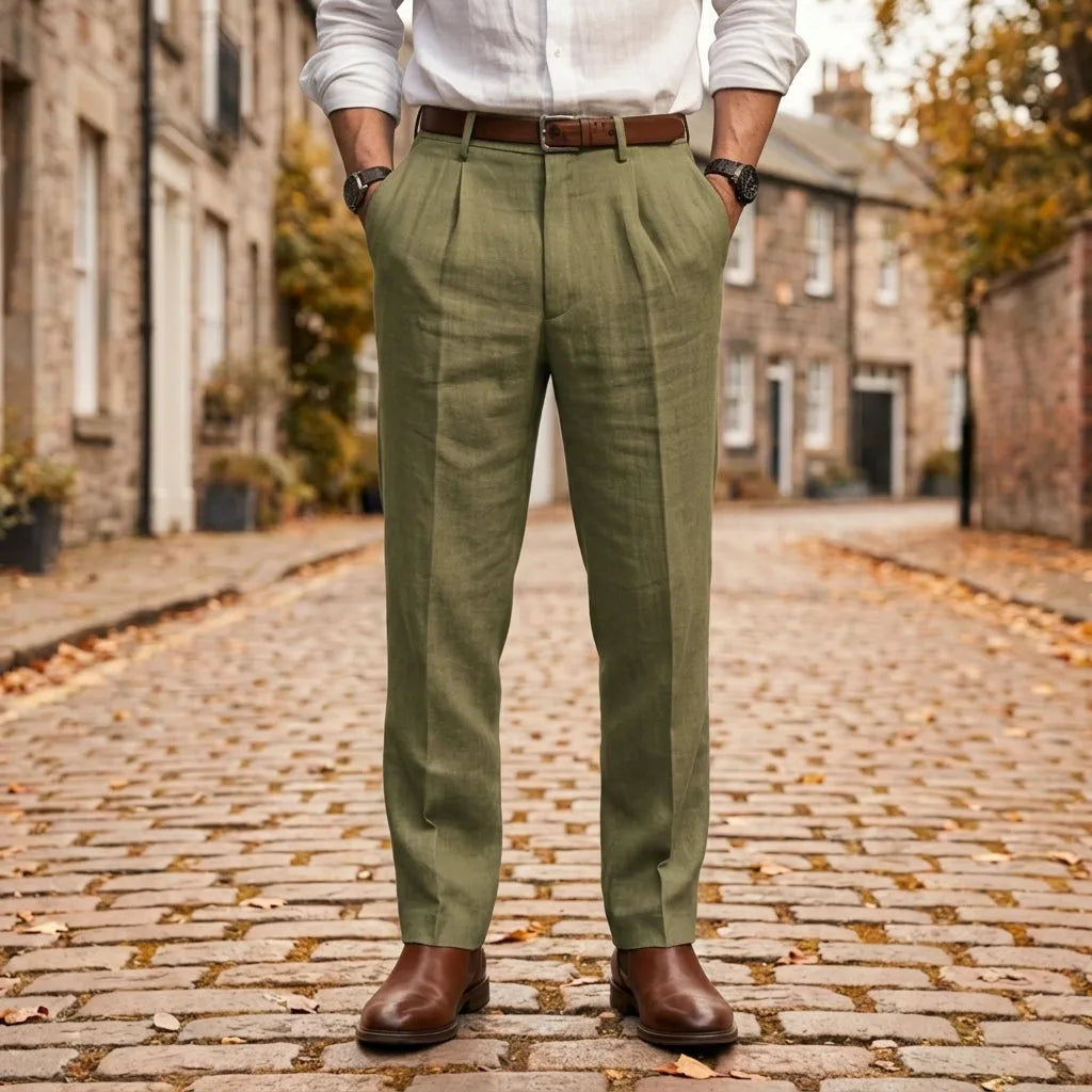 Olive green pleated linen trousers shown with a brown belt and brown Chelsea boots on cobblestones