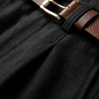 Close-up of the black linen fabric texture, waistband pleat and belt loop with a brown leather belt and brass buckle