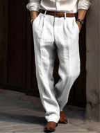 White pleated linen trousers worn with a brown belt, hands resting in the side pockets