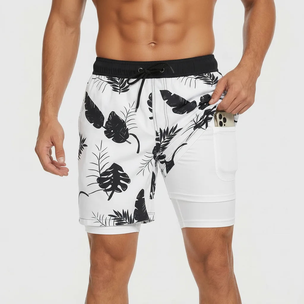 Front view of white swim shorts with black tropical leaf print, pocket pulled open to show white compression liner with phone pocket
