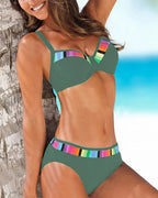 Green two-piece swimsuit with rainbow stripe accents on the bust seam and waistband, shown against a beach backdrop