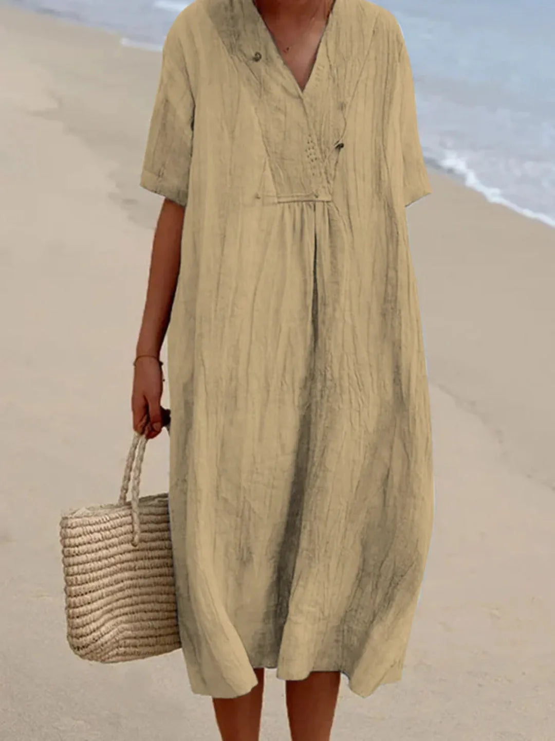 Model in khaki linen-look V-neck midi dress with button detail, carrying a woven straw tote on a sandy beach