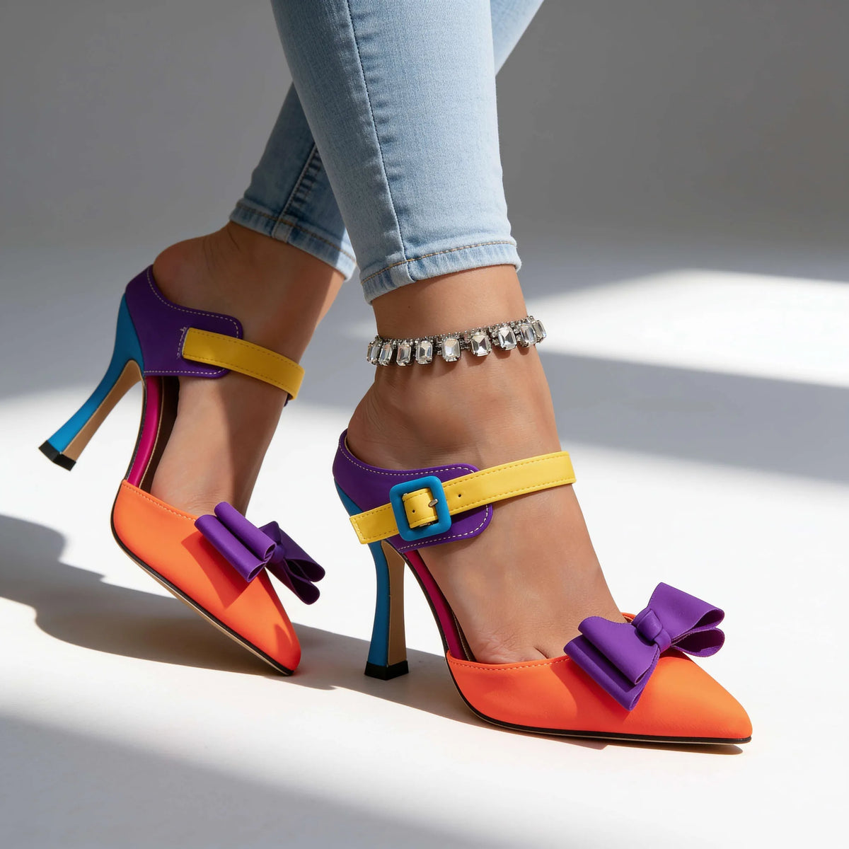 Model walking in orange, purple, yellow and teal stiletto mules against a plain studio background