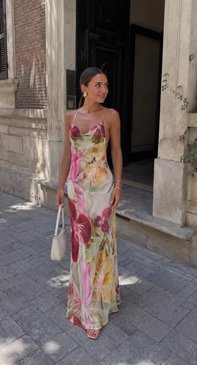 Woman in a rose red floral beaded maxi dress with cowl neckline poses outside an ornate stone doorway, holding a cream handbag